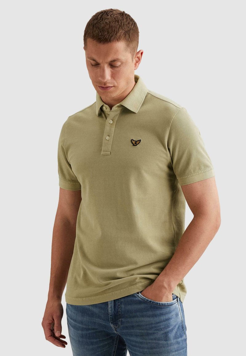 Homme portant un polo vert olive clair avec un petit logo brodé noir et or sur la poitrine, associé à un jean bleu, main dans la poche.