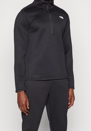 Schwarzer, strukturierter Langarm-Pullover mit Viertelreißverschluss und weißem Logo auf der linken Brust, getragen mit schwarzen Hosen.