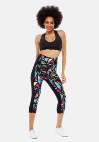 Leggings de capri multicolores con patrones geométricos en tonos brillantes sobre una base negra, combinados con un sujetador deportivo negro y zapatillas blancas.