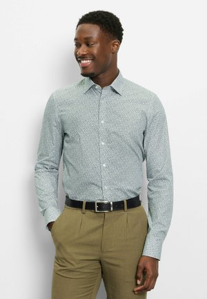 Lächelnder Mann mit kurzen, lockigen Haaren, der ein helles, gemustertes Langarmhemd, khakifarbene Hose und schwarzen Gürtel trägt und mit den Händen in den Taschen steht.