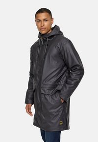 Abrigo gris largo e impermeable con capucha, con cordones ajustables, dos bolsillos frontales y una etiqueta de marca cerca del dobladillo.