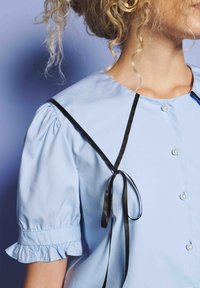 Kvinne iført en lyseblå bluse med puffermer, perleknapper, en bred krage trimmet i svart, og et svart bånd bundet på skulderen.