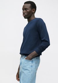 Νέος άντρας φορώντας ένα ναυ蓝 μπλε πουλόβερ και ανοιχτού μπλε τζιν, με το ένα χέρι στην τσέπη, να στέκεται μπροστά σε έναν απλό λευκό τοίχο.