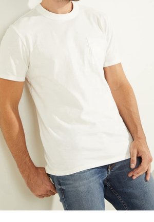 Homme portant un t-shirt blanc à manches courtes avec une poche poitrine et un jean bleu, debout contre un mur blanc, mains détendues le long du corps.