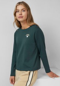 Langsleeve, teal shirt met ribbelnek en geborduurd logo. Zachte stof, losse pasvorm, gecombineerd met beige broek met zijstrepen.