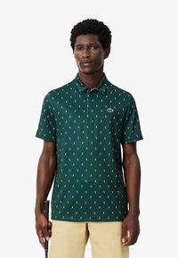 Zielona koszulka polo z powtarzającym się białym wzorem, krótkimi rękawami i kołnierzykiem. Ma małe logo Lacoste na piersi.