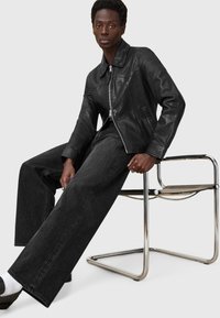 Schwarze Lederjacke mit Reißverschluss, kombiniert mit weit geschnittenen, dunklen Jeans. Das Modell sitzt auf einem schlanken, minimalistischen Stuhl.
