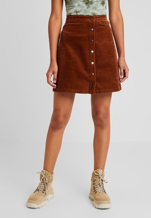 Kobieta w brązowej sztruksowej spódnicy z przodu na guziki, w beżowych sznurowanych botkach, na szarym tle.