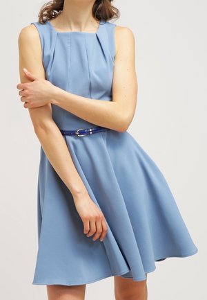 Femme portant une robe bleu clair sans manches avec des plis et une fine ceinture bleu foncé, tenant son bras opposé devant un fond uni.