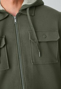 Vyras dėvi alyvuogių žalios spalvos užtrauktuku užsegamą džemperį su šerpa pamušalu, kišenėmis su užsegamomis šoninėmis kišenėmis ir kapisonu su virvute.