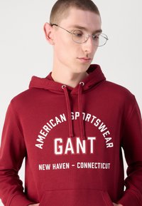 Piros pamut kapucnis pulóver, amely húzózsinórral, elülső zsebbel és fehér nyomtatott szöveggel rendelkezik: "AMERICAN SPORTSWEAR GANT NEW HAVEN - CONNECTICUT."