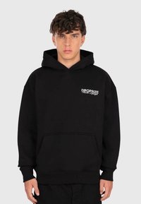Junger Mann mit lockigem Haar, der einen schwarzen Hoodie mit weißem "Dropsize Hustle Lifestyle"-Text auf der Brust trägt und vor einem schlichten Hintergrund steht.