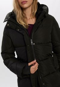 Sort polstret jakke med lynlås og trykknapper, der har hætte, blød inderside og sidelommer. Tekstureret stof med en glat overflade.