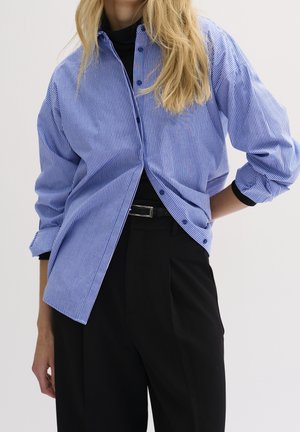 Persona rubia que lleva una camisa de rayas azul y blanca suelta abotonada sobre una camiseta negra, combinada con pantalones negros de tiro alto y un cinturón.