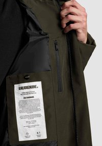 Žalio džemperio vidus su juoda užtrauktuku užsegama kišene, audinio etikete su prekės ženklo duomenimis ir kontrastuojančia blizgia juoda pamušalu.