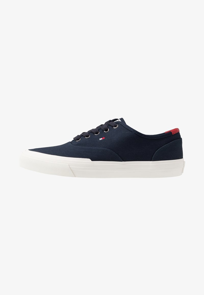 Marineblaue Segeltuch-Sneaker mit weißen Gummisohlen, runder Zehenpartie und Schnürdesign; verfügt über ein kleines rotes und blaues Logo an der Außenseite.