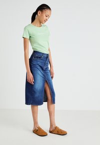 T-shirt vert à manches courtes avec logo blanc, associé à une jupe midi en denim avec une fente sur le devant, et des chaussures slip-on beige.