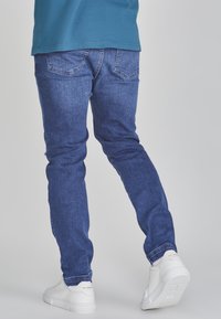 Dark blue denim jeans with a slim fit, featuring back pockets and contrast stitching. Worn with white sneakers on a neutral background.