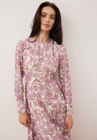 Bloemige jurk van zachte stof met lange mouwen. Kenmerkt zich door een lichtroze en bruine bloemenpatroon, geplooid lijfje en een ronde halslijn.