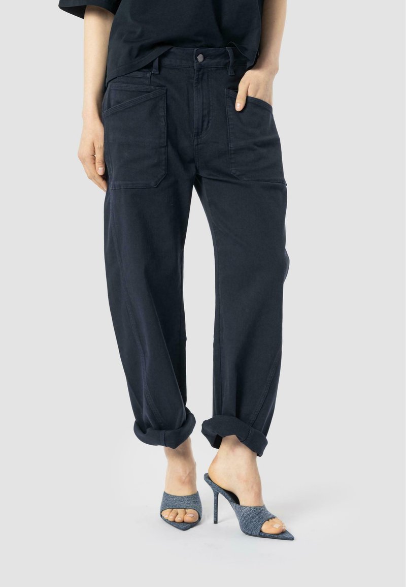Dark wash, loose-fitting denim trousers with rolled cuffs, featuring large front pockets and a button closure. Paired with gray open-toe heels.