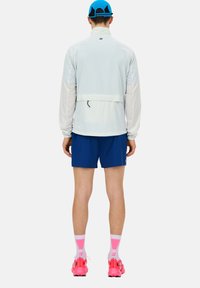 Giacca a vento leggera e traslucida con colletto alto, maniche lunghe e dettagli in rete, abbinata a pantaloni corti blu e scarpe rosa brillante.