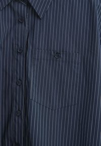 Granatowa, w paski, koszula na guziki z kieszonką na piersi, o gładkiej teksturze i kontrastowych jasnych paskach. Idealna zarówno na co dzień, jak i na formalne okazje.