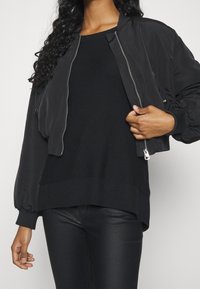 Veste bomber noire avec fermeture éclair à l'avant, poignets côtelés et taille élastique, portée avec un pull noir texturé et un pantalon noir ajusté.