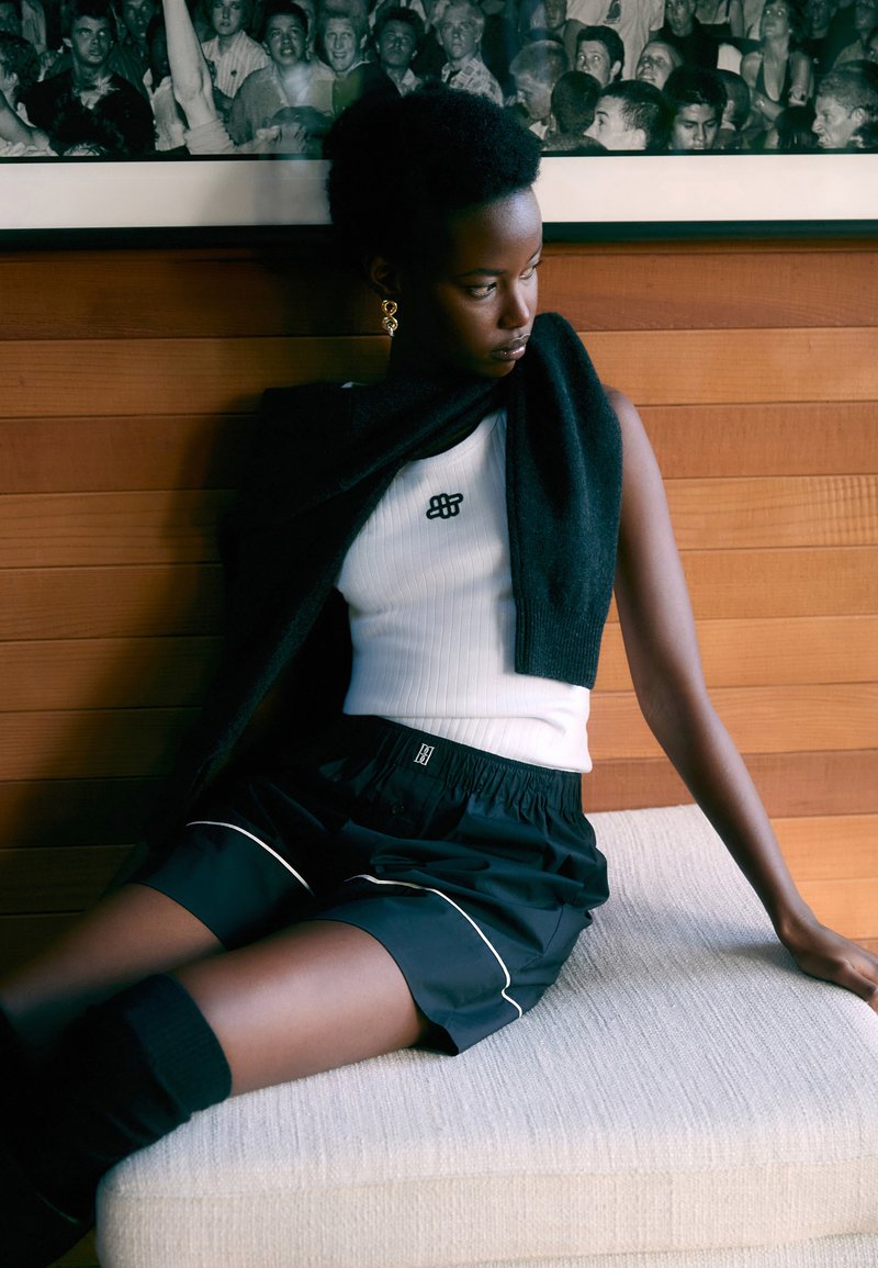 Débardeur noir à côtes avec logo, shorts noirs avec piping blanc, sweat à capuche sombre drapé sur les épaules, assis sur un canapé en tissu texturé clair.