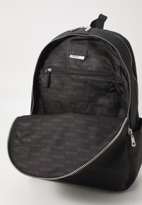 Sac à dos noir avec un extérieur texturé, une fermeture éclair argentée et un logo. L'intérieur présente une doublure à motifs et plusieurs poches de rangement.