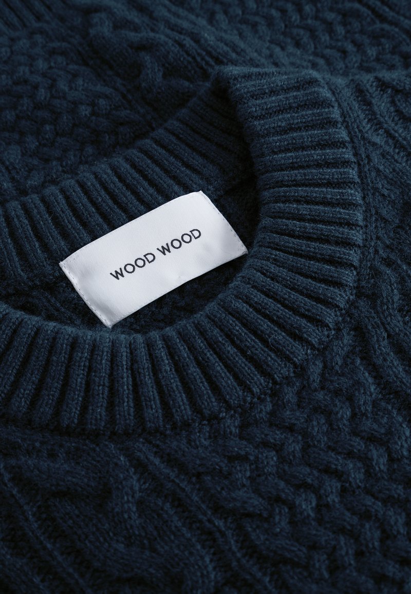 Pull en tricot bleu marine avec des motifs texturés, col rond côtelé et une étiquette blanche affichant la marque "WOOD WOOD."