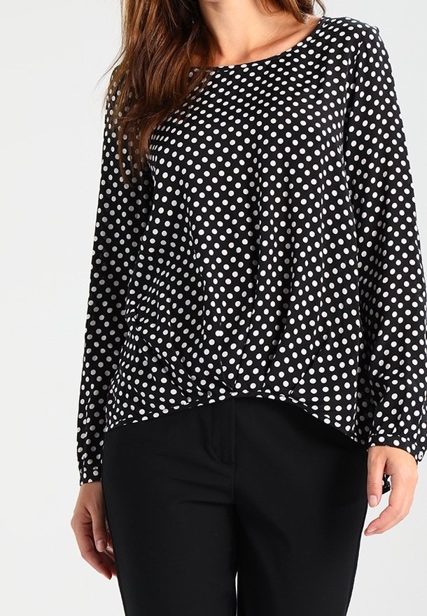 Femme portant un chemisier noir à manches longues avec des pois blancs et un pantalon noir, vue du cou aux hanches.