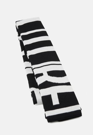 Écharpe tricotée noire et blanche avec de larges rayures horizontales et de grandes lettres blanches partielles sur fond noir, pliée sur une surface blanche.