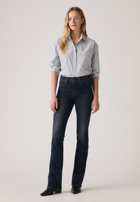 Striped button-down shirt in blue and white, paired with dark denim flared jeans. Model wearing black pointed-toe shoes. Simple background.