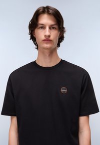 Zwart katoen t-shirt met een rond logo-embleem in oranje en blauw op de linkerborst. Het shirt heeft een klassieke ronde halslijn en korte mouwen.