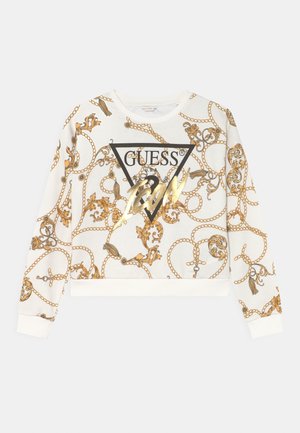 Sweat-shirt blanc à manches longues avec motif chaîne dorée et floral, logo triangulaire noir Guess, et texte "1981" en feuille dorée sur le devant.