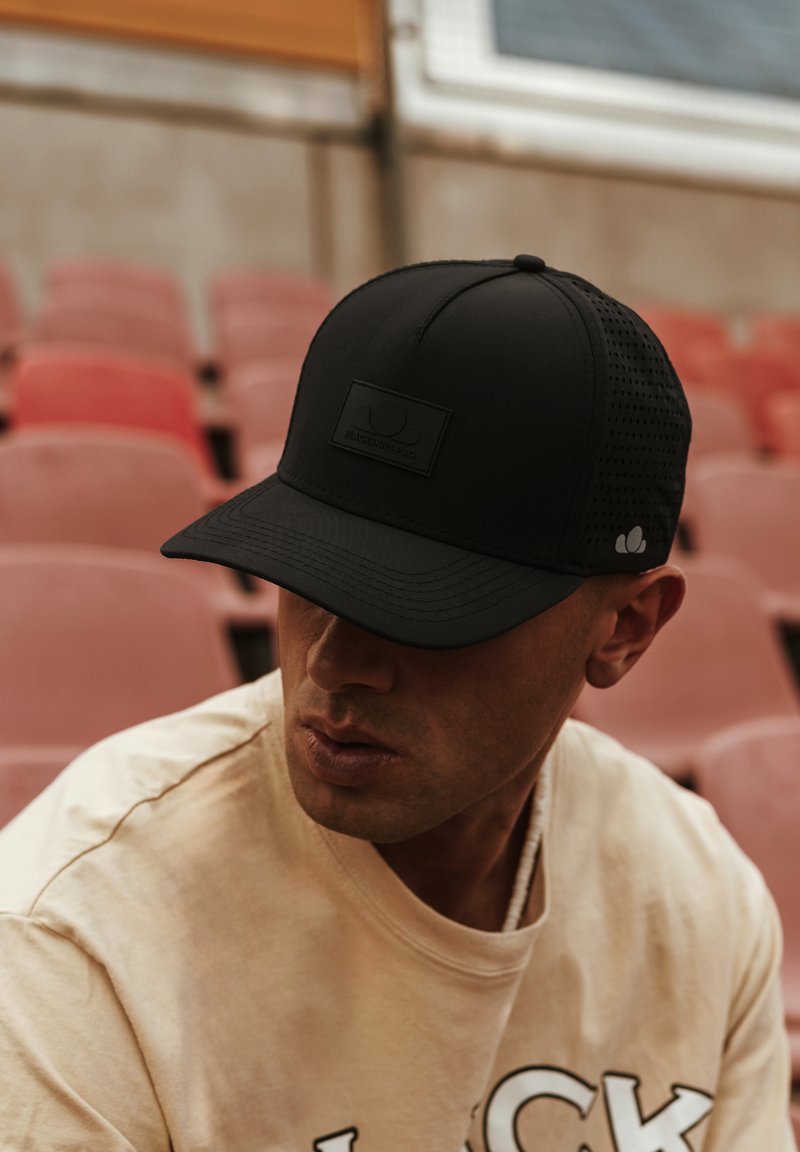 Casquette noire avec une partie en maille à l'arrière, dotée d'un patch texturé, d'un pic arrondi et d'un détail de logo subtil. Fond aux tons neutres avec des sièges vides.