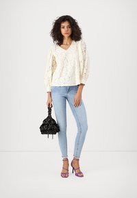 Blusa in pizzo di colore avorio con maniche a sbuffo, abbinata a jeans skinny blu chiaro. Tiene una borsa nera con stampa floreale a cordoncino e indossa sandali stringati viola.