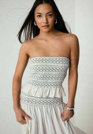 Žena s dlhými tmavými vlasmi v bielych šatách bez ramienok s čiernym prešívaním, striebornými kruhovými náušnicami a strieborným náramkom, stojaca pred jednoduchým pozadím.