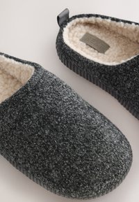 Sivi copati za notranjo uporabo s podlogo iz flisa, z teksturirano zunanjo površino, zaobljenim prstom in črnim gumijastim podplatom s črtami. Vključuje zanko za lažje obuvanje na zadnjem delu.