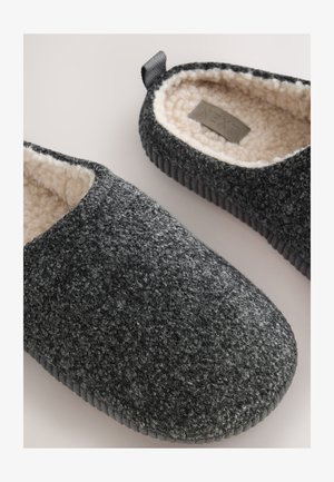 Pantofole grigie foderate in pile con materiale esterno testurizzato, punta arrotondata e suola in gomma nera a strisce. Include un anello posteriore per facilitare la calzata.