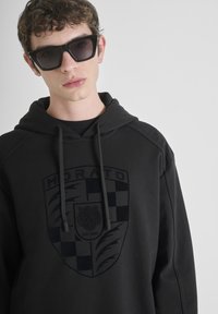 Sudadera negra con capucha, que presenta un emblema tonal en el pecho. La tela es suave y el diseño tiene un corte holgado con cordones.