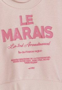 Sweat-shirt rose avec texte en relief "LE MARAIS" et de plus petits mots en écriture cursive et imprimée faisant référence au 3e arrondissement de Paris et à ses lieux culturels.
