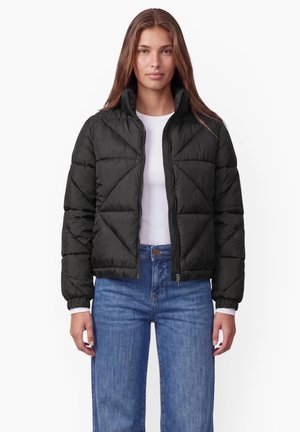 Femme portant une veste matelassée noire sur une chemise blanche et un jean bleu, debout devant un fond blanc.