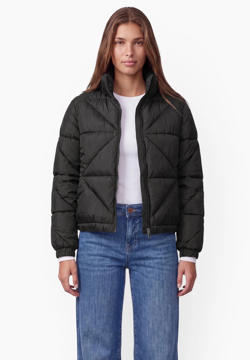 Femme portant une veste matelassée noire sur une chemise blanche et un jean bleu, debout devant un fond blanc.