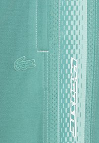 Tissu de sport teal avec une surface texturée, un logo crocodile en caoutchouc et des détails de logo et des coutures blanches contrastantes.