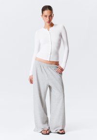 Enganliegende weiße Bluse mit Knopfverschluss und langen Ärmeln, kombiniert mit locker sitzenden, hellgrauen Jogginghosen mit elastischem Bund. Schwarze Sandalen.