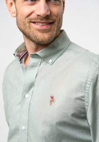 Camicia verde chiaro con bottoni, realizzata in tessuto con una texture liscia. Presenta un piccolo logo ricamato sul petto e un colletto a righe all'interno.