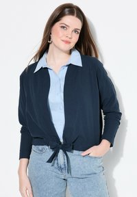 Cardigan navy con chiusura frontale, realizzato in tessuto morbido, con maniche lunghe e vestibilità relaxed, abbinato a una camicia button-up azzurra e jeans.