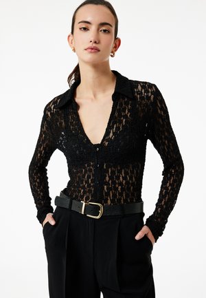 Zwarte kanten overhemd met knopen en een puntige kraag, met lange mouwen. Draagt boven een hoog in de taille gesneden zwarte broek, met een gouden riemgesp.
