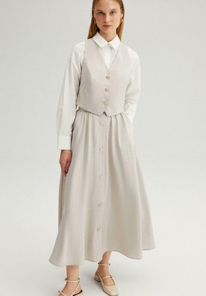 Γυναίκα που φοράει λευκό πουκάμισο με κουμπιά, μπεζ αμάνικο γιλέκο και μακριά φούστα με κουμπιά στο ίδιο χρώμα, στέκεται μπροστά σε έναν απλό φόντο.